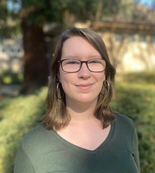 A picture of Anna Boyer: a white woman with shoulder length brown hair, glasses, wearing a green shirt with a nature background