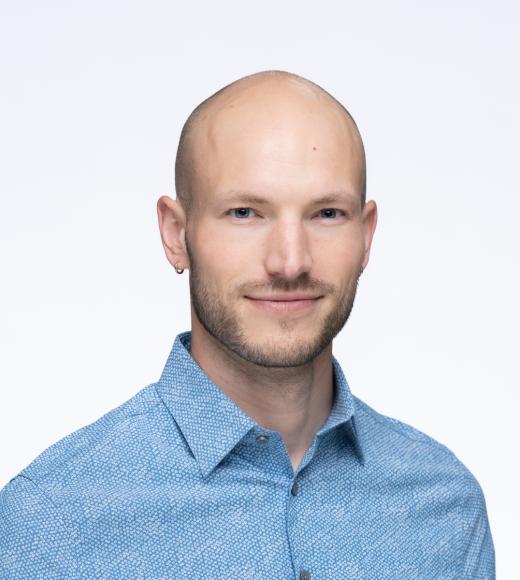 A white bald man in his early thirties, smiling softly and wearing a blue shirt.