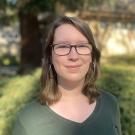 A picture of Anna Boyer: a white woman with shoulder length brown hair, glasses, wearing a green shirt with a nature background