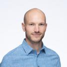 A white bald man in his early thirties, smiling softly and wearing a blue shirt.