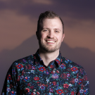 A man smiles in front of a pink and purple sunset with a mountain in the distance. He has white, pale skin, brown hair and a beard, and is wearing a floral red, pink, and purple shirt on a black background. 
